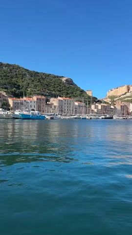 The camera is positioned on a body of water, likely a boat, showing a coastal city with buildings nestled between a verdant hillside and a fortress-like structure perched on the cliffs above. Numerous sailboats are docked in the harbor, creating a picturesque scene under a clear blue sky. The water has a deep blue color with slight ripples on its surface.