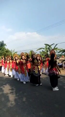 A group of young women dressed in traditional Indonesian attire are walking in a procession down a paved road. They are wearing ornate headdresses, red and gold tops with flowing white skirts or pants, and white shoes. The women are in a line, with some individuals in the front smiling and looking at the camera. The procession appears to be taking place outdoors on a sunny day, with banana trees and other vegetation visible in the background. The sky is clear and blue.
