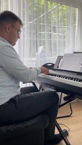 A man wearing a light blue striped dress shirt and dark pants sits at a black digital piano, playing. He has glasses on and is looking at sheet music on a stand. The piano is on a black stand. Behind the piano is a large window covered by white curtains, with green foliage visible outside.