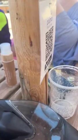 The video opens with a close-up shot of a person's legs and torso, wearing blue and white striped shorts, lying on a beach lounger. A QR code is visible on a sign next to the person, with text reading "PALMAR BEACH BAR SCAN ME TO VIEW MENU". A plastic cup with a straw and some liquid is also on the table. The camera then pans up and out, revealing a beach scene with rows of sun umbrellas and loungers. People are scattered on the beach, some relaxing, and others walking. In the background, there is a beach bar with tables and chairs under a thatched roof and white umbrellas. A young girl in an orange top is standing near the bar. The sky is clear and blue, indicating a sunny day. The video continues to show various views of the beach bar area, with people seated at tables and interacting. The overall atmosphere appears relaxed and typical of a beach resort.
