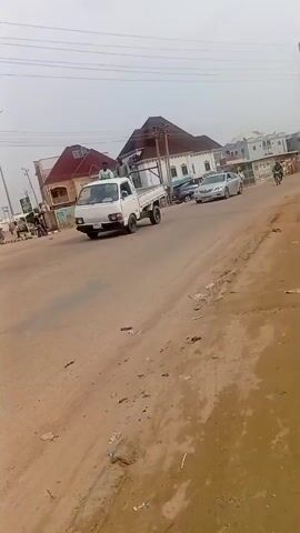 The video shows a street scene in what appears to be a developing country. The road is unpaved and dusty with litter scattered along the sides. Various vehicles including a pickup truck with two people riding in the back, cars, motorcycles, and a three-wheeled vehicle are moving on the road. Buildings are visible in the background, some of which appear to be residential homes. A large orange sign advertising "Study Abroad Made Easy with IDP" is visible on the right side of the road. The sky is overcast and the overall impression is one of a busy street in a developing urban area.