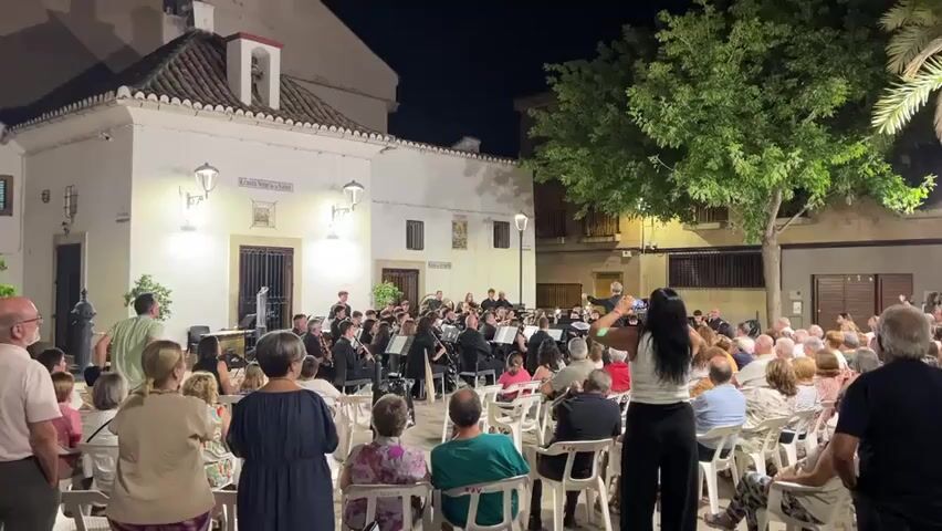 A nighttime outdoor concert features a brass band performing in front of a white building. The band members, dressed in black, are seated on chairs and stand behind music stands. A conductor stands on a platform, leading the musicians. An audience of people of various ages is seated in rows of white chairs, facing the band. Some audience members are visible from behind, with their heads and shoulders in the foreground. The environment includes trees and streetlights. A woman in the foreground, facing away from the camera, is holding up her phone, possibly recording the performance. The scene is illuminated by artificial lighting.