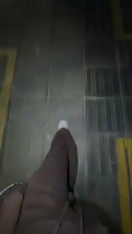 The video begins with a close-up of a person's feet walking on a grated sidewalk. The camera angle shifts upwards, revealing a street scene at night. The sidewalk is paved with square stones and lined with trees. Buildings are visible on the right side of the street, and cars drive on the road. The streetlights illuminate the area. The environment appears wet.
