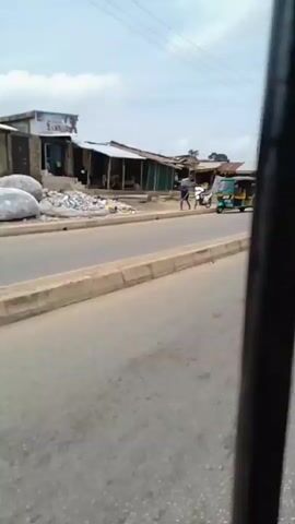 The video shows a street scene in what appears to be a developing area. A paved road with a concrete divider runs through the foreground. On the right side of the road, there are several small, low-lying buildings with corrugated metal roofs, suggesting a commercial or residential area. Some of these buildings have open fronts, possibly serving as shops or stalls. There are a few people visible, and various vehicles including a three-wheeled auto-rickshaw, a motorcycle, and a blue truck. The sky is overcast, indicating a cloudy day. The overall impression is of a busy, somewhat makeshift urban environment.