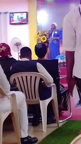 The video shows a scene inside a church. Two women stand on a platform, one leading in singing with a microphone. The platform is decorated with flowers and lights, and there are red stairs leading up to it. An audience sits in chairs facing the platform. A TV screen hangs on the wall, displaying a video. A fan is in the background. There are three people in the foreground, only visible from the back. The environment suggests a religious service or event.