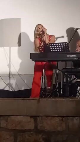 A woman with blonde hair, wearing a red top and red pants, stands center frame, singing into a microphone. She holds the microphone with her right hand and gestures with her left. A black Yamaha keyboard is positioned in front of her on a stand. Behind her and to the right, a woman with long dark hair plays an acoustic guitar. She is wearing a black top. A music stand with a sheet of music is in front of the keyboard. Two microphones are visible, one in front of the singer and one in front of the guitarist. The background is a plain white wall with shadows cast upon it. The scene appears to be a live performance, possibly outdoors, as indicated by the textured stone ledge in the foreground.