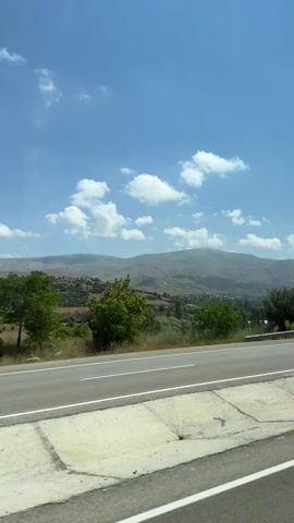 The video shows a view from a vehicle moving alongside a road. The road has two lanes with a white dividing line. A metal guardrail separates the road from a concrete embankment. Beyond the embankment, there is a green landscape with trees and hills. Mountains can be seen in the distance under a bright blue sky with scattered white clouds. The scene appears to be a daytime landscape view during travel.