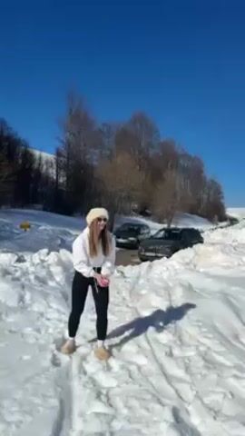 The video shows a woman in a snowy landscape. She wears a white jacket and hat, black leggings, and beige boots. She kicks snow into the air and makes gestures with her hands. She then turns and walks away through the snow. Behind her, there are trees, a snow-covered hill, and two cars parked in the snow. The sky is bright blue.