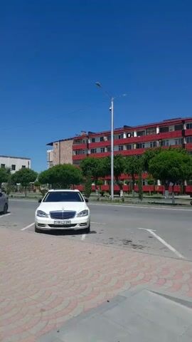 A low-angle, handheld video shows a sunny day in what appears to be a residential area. The video begins with a view of a red apartment building with balconies, across a street. A sidewalk with red and white brick paving is visible in the foreground, and cars are parked along the street. Two people are walking away from the camera on the sidewalk, their backs to the viewer. The person in front is wearing a plaid shirt and dark pants, and the person behind them is wearing a green shirt. Trees are visible along the sidewalk and in front of the apartment building. The video pans slightly to the right, revealing more of the street and sidewalk, and then moves to a more direct view of the sidewalk where people are walking. The video concludes with a view down the street, showing more buildings and trees, and several people walking in the distance. A cell phone tower is visible on top of one of the buildings.