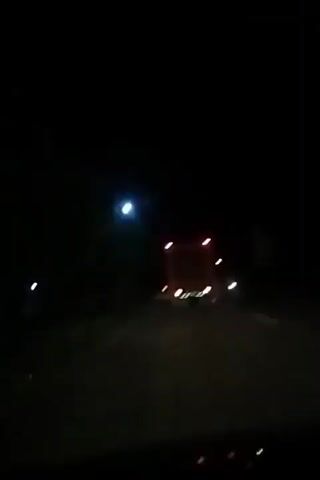 A nighttime video shows a truck driving away from the camera on a dark road. The truck has red taillights and brake lights. There are streetlights visible on the left side of the road. Other vehicles and lights are also visible in the distance. The video quality is low and blurry.