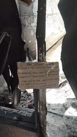 The camera films the interior of a burned house. The walls are dirty, white, and peeling. A sign is posted on a metal frame with the names, phone numbers, and address of Cecilia Cruces Suarez, Sebastian Jara Cruces, and Valentina Jara Cruces. The last line of the address says, “Sea la madre de Jehova bendita?” The floors are covered with debris from the fire. The camera pans around the interior of the house.