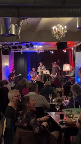 The scene depicts a jazz band performing live in a cozy indoor venue. The band consists of six members: a guitarist, a drummer, a saxophonist, a bassist, and a pianist. They are positioned on a stage adorned with black curtains and lit with blue and red stage lighting. Two chandeliers hang above the band. The audience is seated at tables, enjoying drinks and watching the performance. Most of the audience members are facing the stage, with a few looking to the side or conversing. There are glasses of wine and beverages on the tables, and the overall ambiance is relaxed and sophisticated. Some individuals appear to be using their phones. A pillar with a blue light fixture stands on the left side of the stage.