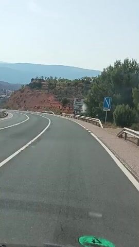 The video is shot from the perspective of someone inside a vehicle, likely a car or van, as it drives along a paved road. The road curves to the left and then to the right, with a white line marking the center and a dotted white line marking the edge on the left. Guardrails run along the right side of the road. On the left, a hillside slopes upwards, dotted with some trees and houses, and in the distance, mountains are visible under a clear sky. On the right, the hillside is also visible, with more sparse vegetation and some red soil. A road sign with destinations such as TORREBAJA, TERUEL, and CASTILLODEFLORES is visible on the right side of the road. The driver's side of the windshield is visible at the bottom of the frame, with a green sticker. The video appears to be taken during the daytime.