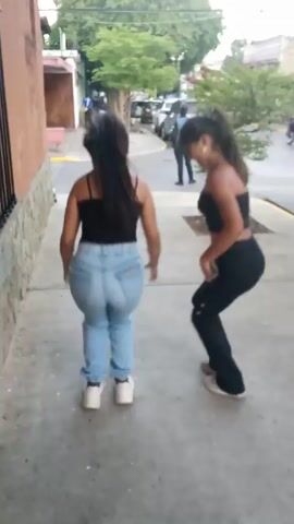 Two young women are seen walking away from the camera on a sidewalk next to a building. The woman on the left has long, dark hair and is wearing a black tank top and light-wash jeans. The woman on the right also has long, dark hair tied in a ponytail and is wearing a black tube top and black pants. They are initially seen holding hands, and then proceed to walk, alternating between skipping and walking. In the background, there are parked cars, a tree, and people standing on the street. The building adjacent to the sidewalk is a peach color.