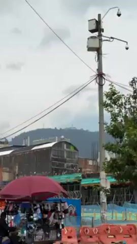 The video shows a tall white church with a pointed steeple atop a hill shrouded in haze. In the foreground, part of a building under construction is visible, with workers in yellow hard hats moving about. A red umbrella and a utility pole with cables are also in view, against a cloudy sky. The camera zooms in slightly to focus on the church.