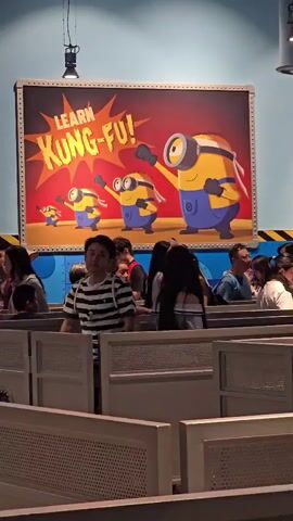 The video shows a queue of people waiting in what appears to be an indoor amusement park attraction. The walls are painted a light blue, and there are yellow and black hazard stripes along the bottom of the wall. Dominating the background is a large, rectangular sign featuring cartoon Minions from the Despicable Me franchise. The sign has a red background with a yellow starburst effect and reads "LEARN KUNG-FU!" in large white letters. Three Minions are depicted on the sign, two in mid-action poses with their fists raised, and one larger Minion at the front also in a dynamic pose. The people in the queue are facing towards the sign, or are in profile. They are mostly looking forward, presumably towards the attraction they are queuing for. Some are in casual clothing, and some are carrying bags. The queue is divided into sections by white, slatted barriers. The lighting appears to be artificial, with a spotlight visible hanging from the ceiling above the sign. The overall impression is of a family-friendly attraction.