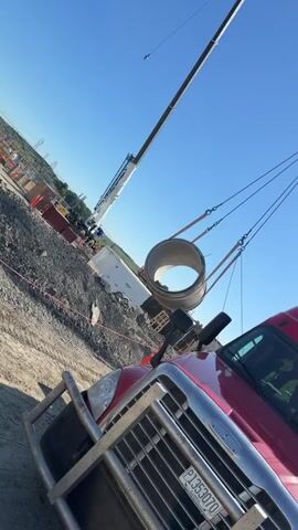 A large white Liebherr crane, positioned on a construction site in Center Township, United States of America, is actively lifting a substantial cylindrical concrete pipe. The pipe is suspended by multiple slings and chains connected to the crane's hook block. The front end of a red semi-truck is visible in the immediate foreground, with part of its license plate reading "P135307". The ground is rugged, covered in dirt and gravel, indicative of a work-in-progress construction area. In the background, under a clear blue sky, other construction materials, a white trailer, and utility poles can be seen. The scene is illuminated by bright daylight, suggesting either morning or afternoon, with no clouds visible. No people are clearly visible, and therefore no emotions can be discerned.