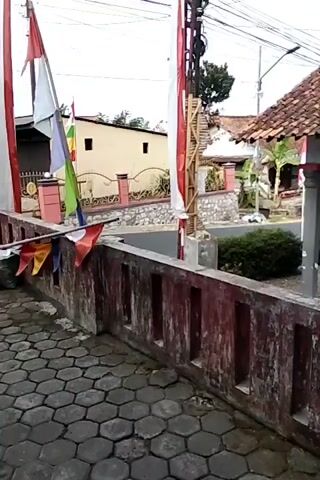 The video shows an outdoor scene with several flags, including Indonesian flags, prominently displayed. The flags are mounted on poles and strung across a railing. The environment appears to be a residential area, with houses and a street visible in the background. The railing itself is made of concrete and has a red, weathered appearance. The ground in the foreground is paved with hexagonal tiles. The video is short and seems to be a static shot with no significant movement.