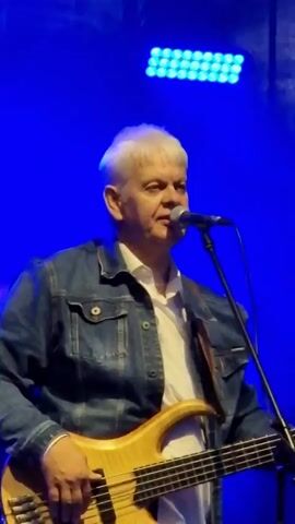 A medium shot shows a white-haired man with a denim jacket over a white collared shirt, playing a light-colored bass guitar and singing into a microphone. He is on a stage with a blue backdrop and a horizontal line of blue lights above him. The man's left hand is on the fretboard of the guitar, and his right hand is near the pickups. He is looking slightly down and to his right as he sings. The video is silent.