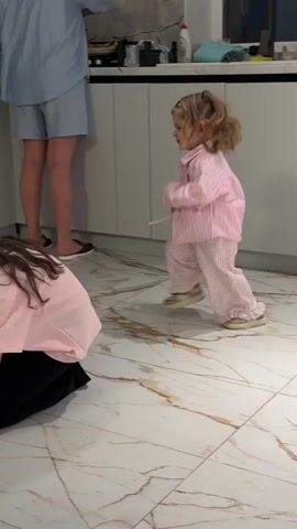 A young girl in pink striped pajamas jumps and skips around a kitchen with a marble floor. She turns towards the camera and screams with an angry expression. She is standing behind a gray metal chair. Another person is partially visible in the foreground.