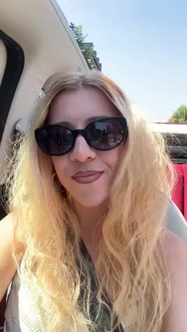 The video begins with a close-up shot of a blonde woman with long, wavy hair wearing black sunglasses and dark lipstick. She is in the passenger seat of a car, with a white seatbelt visible across her chest. The background shows buildings with blue and pink facades, suggesting an urban environment. The woman is smiling with her mouth open, appearing to be singing or speaking excitedly. As the video progresses, she raises her right arm and moves her hand upwards and outwards. The car appears to be in motion, as indicated by the slight blur of the background and the woman's hair blowing in the wind. The sun is shining brightly, casting shadows and highlighting the woman's features. The overall tone of the video is energetic and joyful.