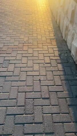 The video begins with a close-up of a cobblestone path. Sunlight streams in from the right side of the frame, casting a golden glow and long shadows. As the camera pans upward, the sun becomes the dominant feature, a bright, almost blinding orb low in the sky. In the background, a street is visible with buildings on either side. One building has a sign that reads "АПТЕКА" (Apteka), which is Russian for pharmacy. A street lamp stands tall, its silhouette against the bright sun. The scene appears to be an outdoor public space, possibly a town square or a pedestrian walkway, bathed in the warm light of either sunrise or sunset. There are no people visible in the video.