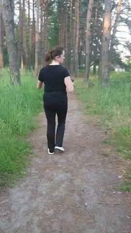 A woman is captured in a video walking along a dirt path through a forest. She is wearing a black t-shirt, black pants, and white sneakers. The path is lined with tall, slender pine trees and lush green grass. The lighting suggests it is either early morning or late afternoon, with soft sunlight filtering through the trees. The woman appears to be looking over her shoulder, perhaps at someone or something behind her. There is no visible text in the video. The overall scene conveys a peaceful and natural setting, likely a park or a well-maintained trail within a wooded area.