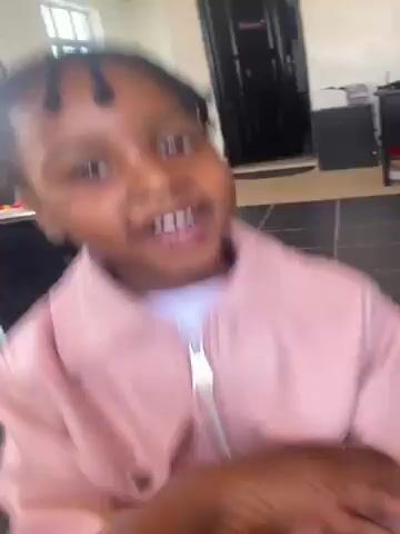A young child with braided hair is wearing a pink jacket and sticking their tongue out. The child is indoors in a living room area with a couch and other children visible in the background. The child moves around and looks at the camera and to the side.