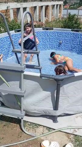 This video shows four young girls playing in a backyard swimming pool. The pool is circular with a blue mosaic tile pattern inside and a gray outer wall. A silver ladder with gray steps leads into the pool. The girls are wearing swimsuits and appear to be enjoying themselves. 

One girl, wearing a pink and white striped swimsuit, is standing on the ladder, looking down into the pool. Another girl, in a pink and white polka dot swimsuit, is lying on her stomach in the water, with her head turned to the side. A third girl, in a colorful swimsuit, is lying on her back with goggles on her eyes, her legs bent. The fourth girl, in a pink and white bikini, is partially visible at the bottom left of the frame, seemingly reaching for something in the water.

The scene is set outdoors on a sunny day, suggested by the bright light and clear blue sky. In the background, there are some unfinished brick structures, a fence, and green vegetation. Distant hills or mountains are visible under the bright sky. The overall atmosphere seems to be one of fun and recreation on a summer day. There is no visible text in the video.