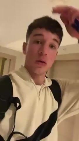 A young man with short, curly brown hair, light skin, and green eyes is filming a close-up selfie video. He is wearing a cream-colored hoodie with black wavy stripes and a black backpack. He is walking through a hallway with light-colored walls and flooring. There are signs visible on the wall behind him. His expression is neutral. He looks directly at the camera. The video appears to be taken indoors.