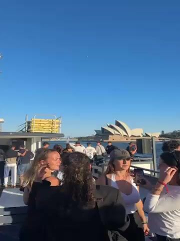 The video shows a group of people on a boat, seemingly a party boat, with the Sydney Opera House in the background. The sky is clear and blue. People are standing and talking amongst themselves. Some are looking towards the Opera House. There is music playing. A yellow structure is on the front of the boat.