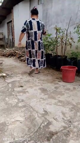 A woman in a patterned dress is watering plants. She is holding a watering can and pouring water onto the plants. The plants are in black bags and are arranged in a row. There is a red bucket next to the plants. The woman is standing on a concrete path. The background shows a building and a pile of wood.