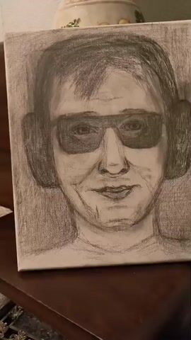A close-up, medium shot, from a slightly low angle, shows a pencil sketch on canvas of a person wearing large headphones and sunglasses. The drawing depicts the upper chest, shoulders, and head of the person. The background behind the canvas is out of focus, but appears to be a room with furniture. The sketch is done in shades of gray and black with some areas showing the texture of the canvas. The person in the sketch has dark hair, a neutral facial expression with a slight smile, and appears to be looking directly at the viewer. The video pans slowly from left to right across the canvas.