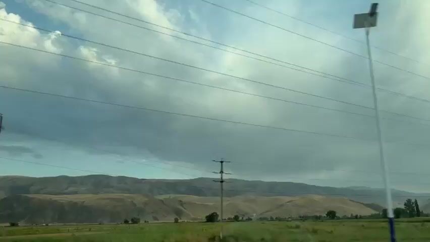 The video shows a landscape from a moving vehicle. The sky is cloudy with patches of blue. There are several power lines crisscrossing the frame. In the foreground, a white pole with a black light fixture is visible on the left. Further in the distance, there are rolling hills and some trees. The vehicle appears to be moving along a road, as indicated by the passing scenery and the motion blur. The overall impression is one of a rural or semi-rural environment under a dynamic sky.