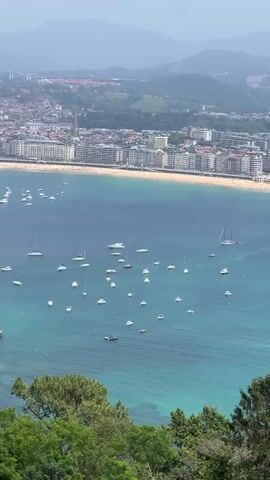 A high-angle shot shows a scenic view of a bay filled with boats. The turquoise water is dotted with numerous sailboats and smaller vessels. In the background, a curved beach is lined with buildings and a promenade, suggesting a coastal city. The cityscape extends inland, backed by hazy green hills. Lush green trees frame the bottom of the shot, adding to the natural beauty of the scene. The sky is overcast, casting a soft light over the entire landscape. There are no discernible people in the water or on the beach.