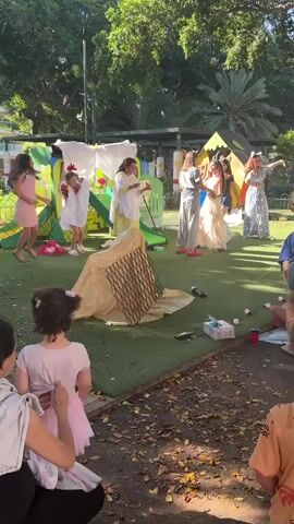 The video shows a group of children and adults outdoors on a grassy area. Several children are dressed in costumes, some appear to be performing or dancing on a makeshift stage with colorful backdrops. One child in the foreground, facing away from the camera, wears a pink tutu and is watching the performance. Another child, also facing away, is dressed in an orange t-shirt. There are scattered objects on the ground, including a gift-wrapped box and what appear to be small white balls. Trees and foliage are visible in the background, suggesting a park or outdoor recreational area. The lighting suggests it is daytime, likely late afternoon due to the warm, dappled sunlight.
