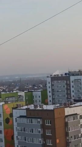 The video shows an urban landscape with several multi-story buildings, viewed from an elevated position, likely from another building. The buildings vary in color and design, some with decorative patterns on their facades. In the background, there is a hazy view of more buildings and perhaps some industrial structures in the distance. The sky is pale and overcast. A wire stretches across the upper part of the frame.