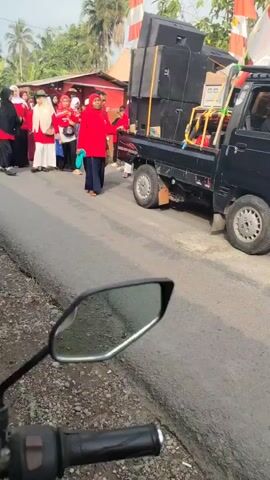 A black pickup truck is loaded with speakers and audio equipment. Several people, some wearing red shirts and head coverings, are walking alongside the truck in a street. The truck is moving slowly, and the people appear to be part of a procession or parade. The scene is outdoors, and there are flags and buildings visible in the background. A motorcycle's rearview mirror and handlebars are in the foreground, suggesting the video is being recorded from another motorcycle.