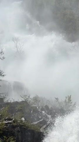 The video shows a close-up view of a waterfall cascading over rocks and vegetation. The water appears white and foamy as it crashes down. Mist or fog fills the air, obscuring the surrounding environment. The rocks are dark and partially covered in green plants. Bare tree branches are visible in the background.