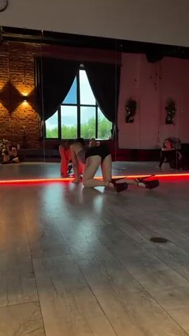 In a studio with a wooden floor and a large mirror reflecting the scene, two individuals are engaged in a dance or movement practice. The person in the foreground is on their hands and knees, wearing black shorts, a gray top, and high-heeled shoes with red soles. Their back is to the camera, with tattoos visible on their back and leg. The mirror shows another person in a similar pose, facing the first individual. The studio has exposed brick walls and large windows with dark curtains, suggesting an indoor setting. Red LED lights line the base of the mirror, casting a glow on the floor. The overall atmosphere appears to be focused and possibly sensual, given the attire and poses. The time of day is not explicitly evident, but the lighting suggests it could be daytime or early evening. The location is identified as Vladimir, Russia.