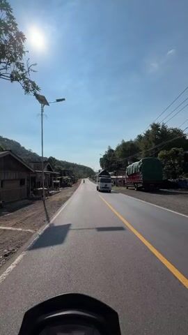 A first-person perspective video shows a motorcycle traveling down a paved road under a clear blue sky. The sun is bright and high in the sky, casting a glare. On the left side of the road, there are several small buildings, some with awnings and open stalls. A solar-powered street lamp stands prominently on the left. On the right side of the road, there are more buildings, some covered with green tarps, and parked motorcycles. Two trucks are visible on the right side of the road, one white and one red, also covered with tarps. A single person is seen riding a motorcycle in the distance ahead, and another individual on a motorcycle is further ahead, approaching the viewer. The road has a single yellow line marking the center. The motorcycle's dashboard displays a speed of 55. The surrounding environment appears to be a rural or village setting with some vegetation and hills in the background.