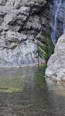 The video shows a narrow canyon with a waterfall cascading down a rocky cliff face. The water flows into a clear pool at the bottom. The canyon walls are made of grey rock with patches of green moss growing on them. The water is clear and reflects the surrounding rocks and vegetation. The camera pans upwards to follow the waterfall.