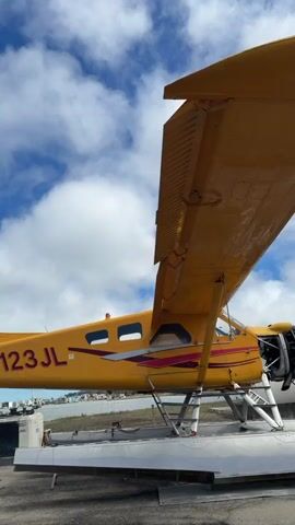 A close-up, low-angle shot shows the side of a yellow seaplane with the registration number N123JL visible on the fuselage. The plane is equipped with floats and rests on a tarmac or dock. The sky above is blue with scattered white clouds. The scene appears to be outdoors, likely at an airport or marina, with a body of water and distant structures visible in the background.