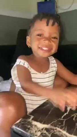 A young child, probably a toddler, is seen in the video. She is wearing a striped white and grey tank top, has dark skin, and is wearing small hoop earrings. Her hair is styled in braids. The child is sitting on someone's lap, initially at a table, and then laid back onto a black leather couch. The child is crying, looking up, and moving their hands. A person's legs are visible, wearing light-colored shorts. In the background, there is a dark-colored couch and a blue object on a shelf or table. The lighting in the room is moderate.