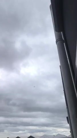 The video shows a cloudy sky with a small airplane flying in the distance. The camera is angled upwards, showing the underside of a building with a pipe running down the side. In the background, there are some buildings and a body of water. The scene is overcast and appears to be daytime.