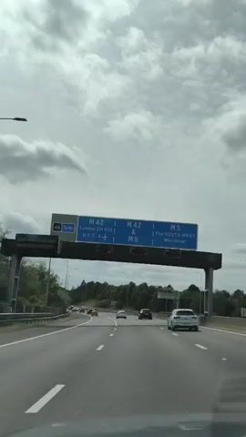 The video shows a POV shot from inside a car driving on a highway. The car is in the right lane, with other cars visible ahead and to the left. Overhead, a large blue highway sign indicates directions: "M42 & M5" for London (M40), N.E.C. & ✈️, and "THE SOUTH WEST" & Worcester. The sky is overcast with clouds, and trees line the side of the highway. The road has white dashed lines marking the lanes. The timestamp indicates the video is a few seconds long, showing continuous forward motion on the highway.