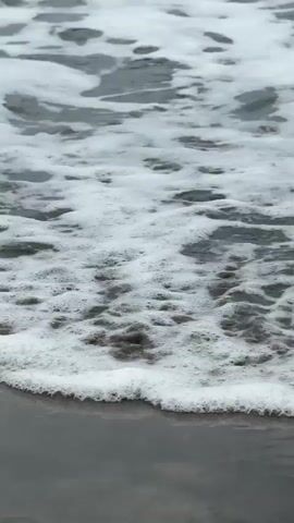 A close-up shot focuses on the surface of the ocean. The water is grey and choppy, with white foam creating swirling patterns. The camera is positioned at a downward angle, showing the foam and water moving towards the viewer. There are no people or other objects visible in the frame. The overall impression is of a dynamic and turbulent body of water.