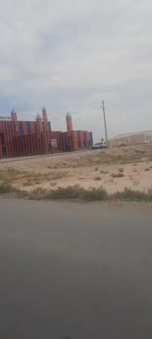 The video shows a building with orange and blue striped walls and several towers. The building has the text "SEN 7 SEN" on it. There are several cars parked in front of the building, including a white SUV and a silver sedan. The area around the building is dry and dusty with some sparse vegetation. The sky is cloudy.