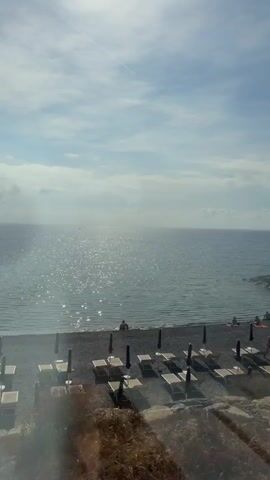 The video shows a view of a beach on a sunny day. The water is calm and reflects the sunlight. There are several people on the beach, some sunbathing, others walking or swimming. There are also beach chairs and umbrellas set up on the sand. In the foreground, there are some rocks and foliage. The video pans slowly across the scene, offering a glimpse of the beach and the sea.