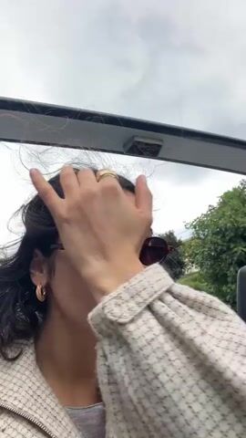 A woman takes a selfie-style video while riding in the backseat of an open-air vehicle, possibly a bus or jeep, with a man seated next to her. The woman has dark hair, fair skin, and is wearing brown sunglasses, gold hoop earrings, a ring, and a white speckled sweater. She looks at the camera, adjusts her sunglasses, and puts her hand behind her head. The man next to her has curly dark hair and is wearing black sunglasses and a black leather jacket. Outside the vehicle, greenery and road signs are visible, suggesting they are driving in a semi-rural area on an overcast day. The camera angle is slightly upward, showing the vehicle's open roof. The overall mood seems relaxed and casual. Music is playing in the background.